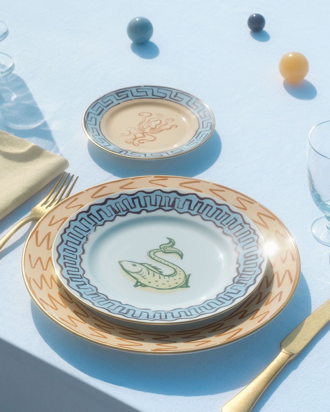 Stacked decorative ceramic plates with green fish motif and Greek‑key border on a pale blue tablecloth, with gold cutlery, glass and coloured marbles