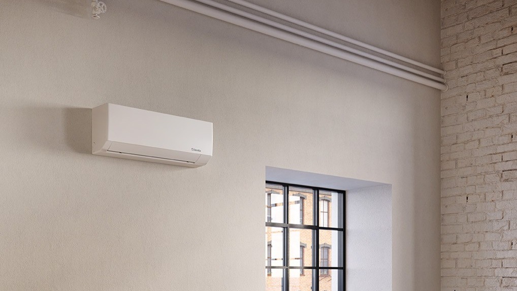 Wall-mounted white air conditioner on a pale plaster wall in a loft-style room with industrial window and exposed brick