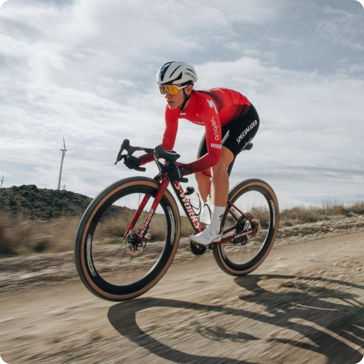 Cyclist in red kit and aero helmet racing a red road bike on a gravel trail with wind turbines under a cloudy sky