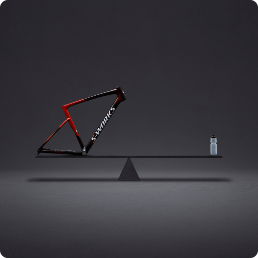 S-Works red and black carbon road bike frame balanced on a seesaw opposite a water bottle against a minimalist dark background