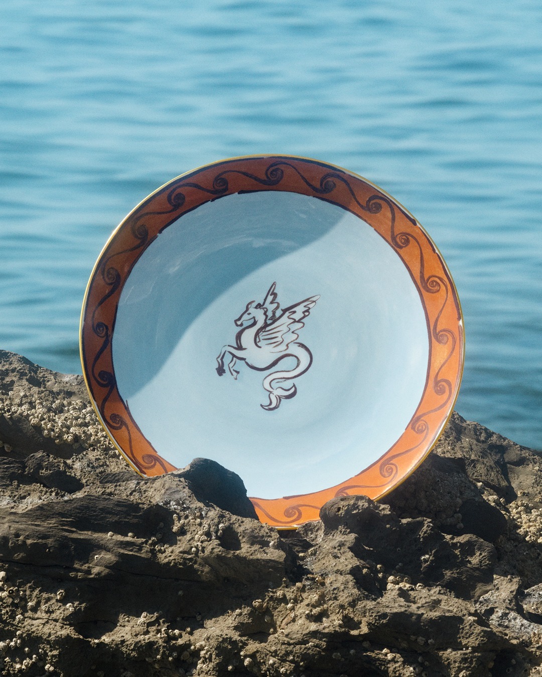 Ceramic plate with orange decorative rim and central winged horse (Pegasus) motif, propped on rocky shore with sea background