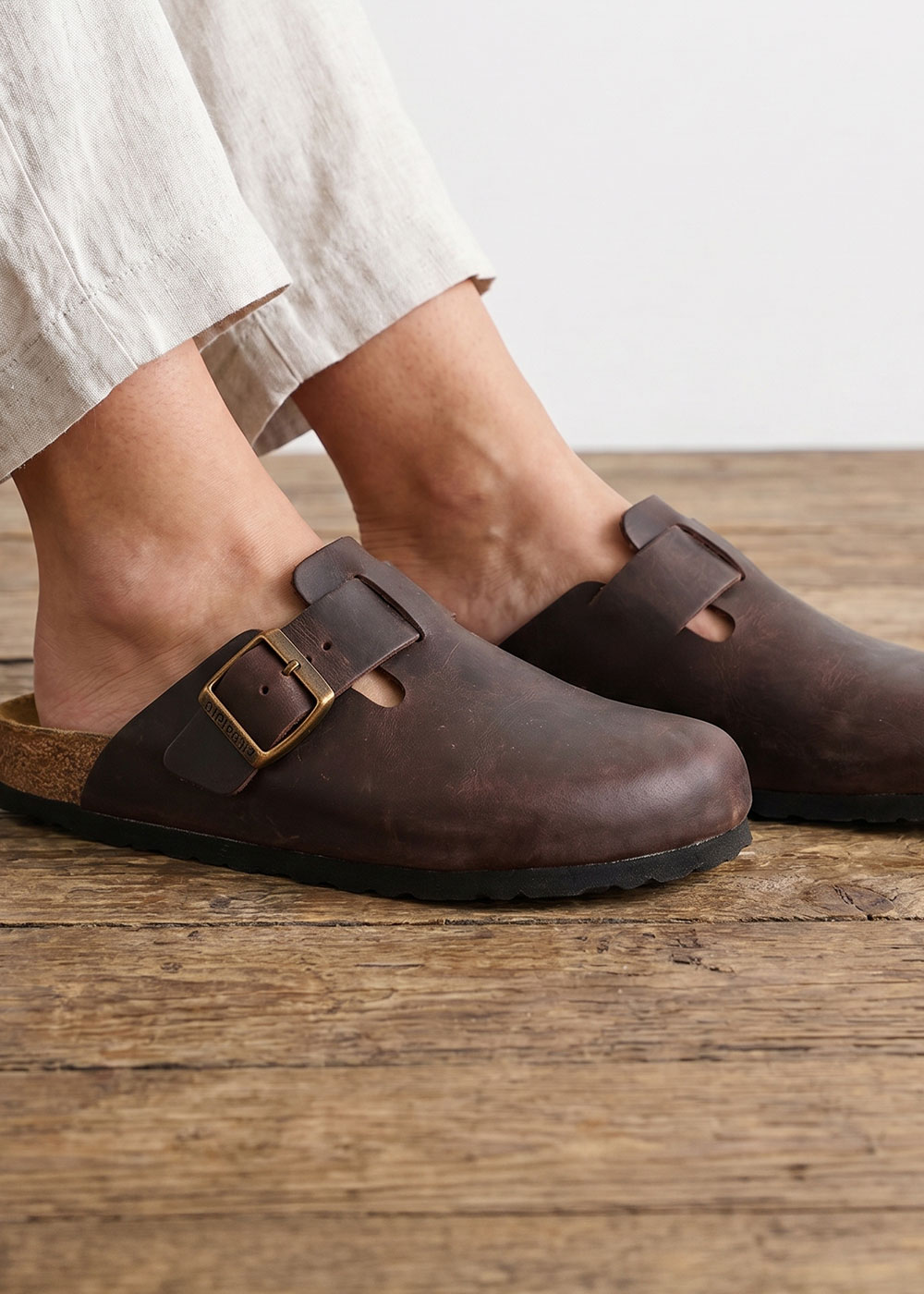 Brown leather slip-on clogs with brass buckle and cork footbed on wooden floor, worn with cropped linen trousers
