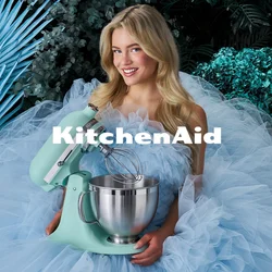 Donna in un voluminoso abito di tulle azzurro chiaro che tiene un mixer da tavolo verde menta, circondata da foglie blu-verdi e cupcake su un piccolo tavolo.
