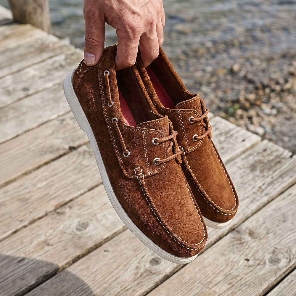 Hand holding brown suede boat shoes with white soles above a weathered wooden dock and pebbled shoreline