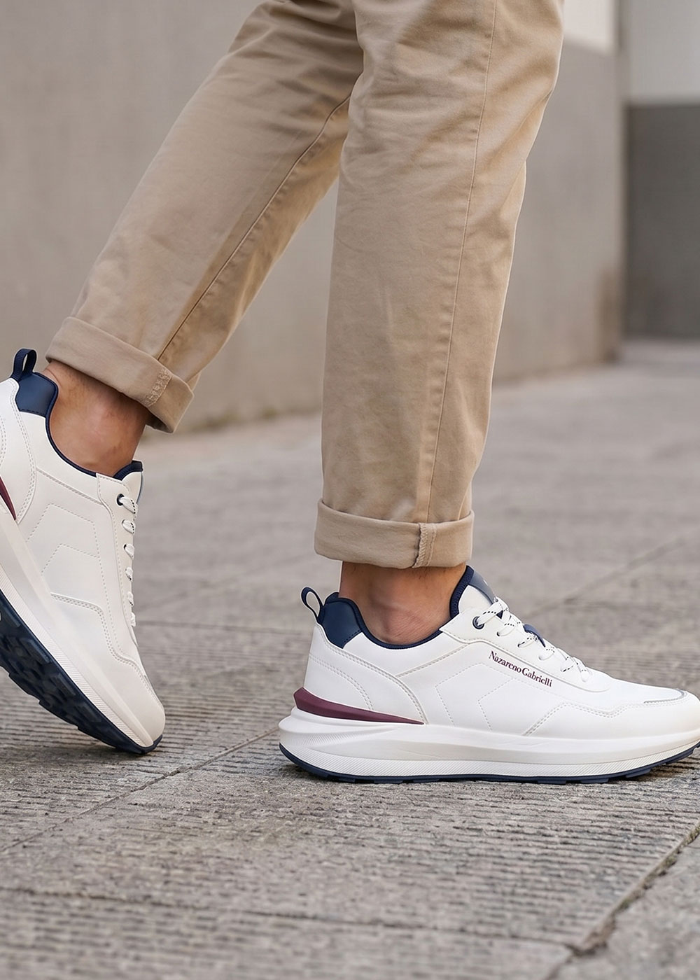 Close-up of a person walking on pavement wearing white trainers with navy and maroon accents and rolled-up beige chinos