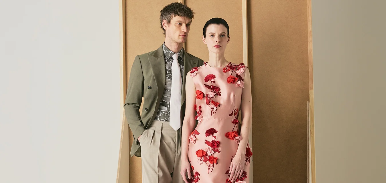 Uomo con blazer verde oliva e pantaloni beige sta accanto a una donna con un vestito floreale rosa su uno sfondo beige minimalista.
