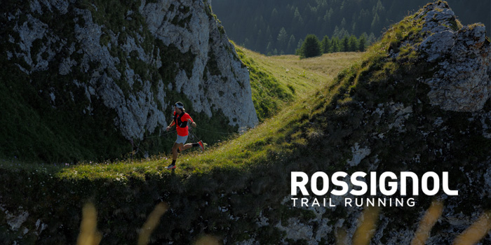 Trail runner in red jacket on a narrow mountain ridge with rocky cliffs and forested valley, Rossignol Trail Running logo