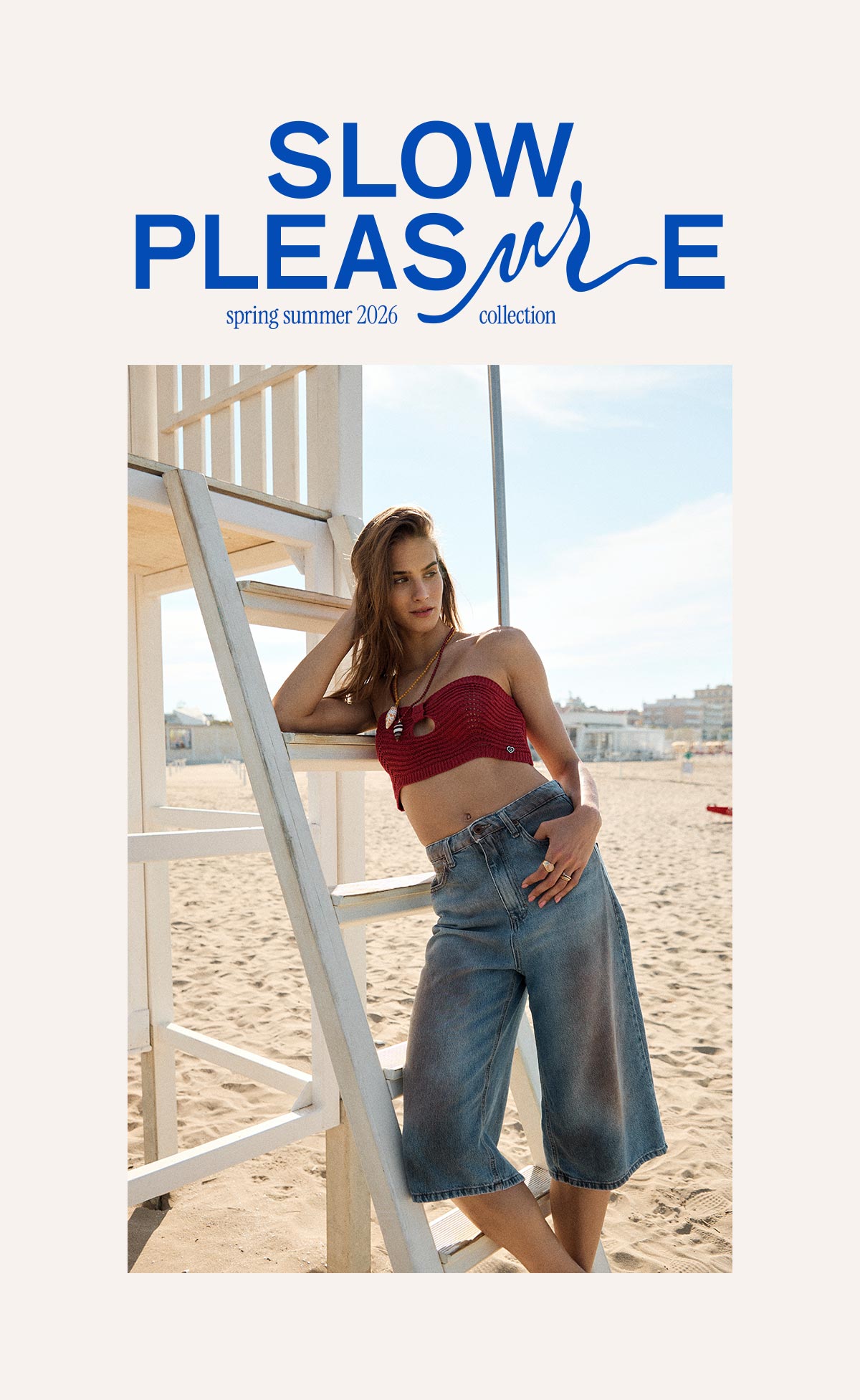 Model leaning on lifeguard tower at sunny beach wearing red knitted crop top and wide‑leg denim culottes, SLOW PLEASURE SS26.