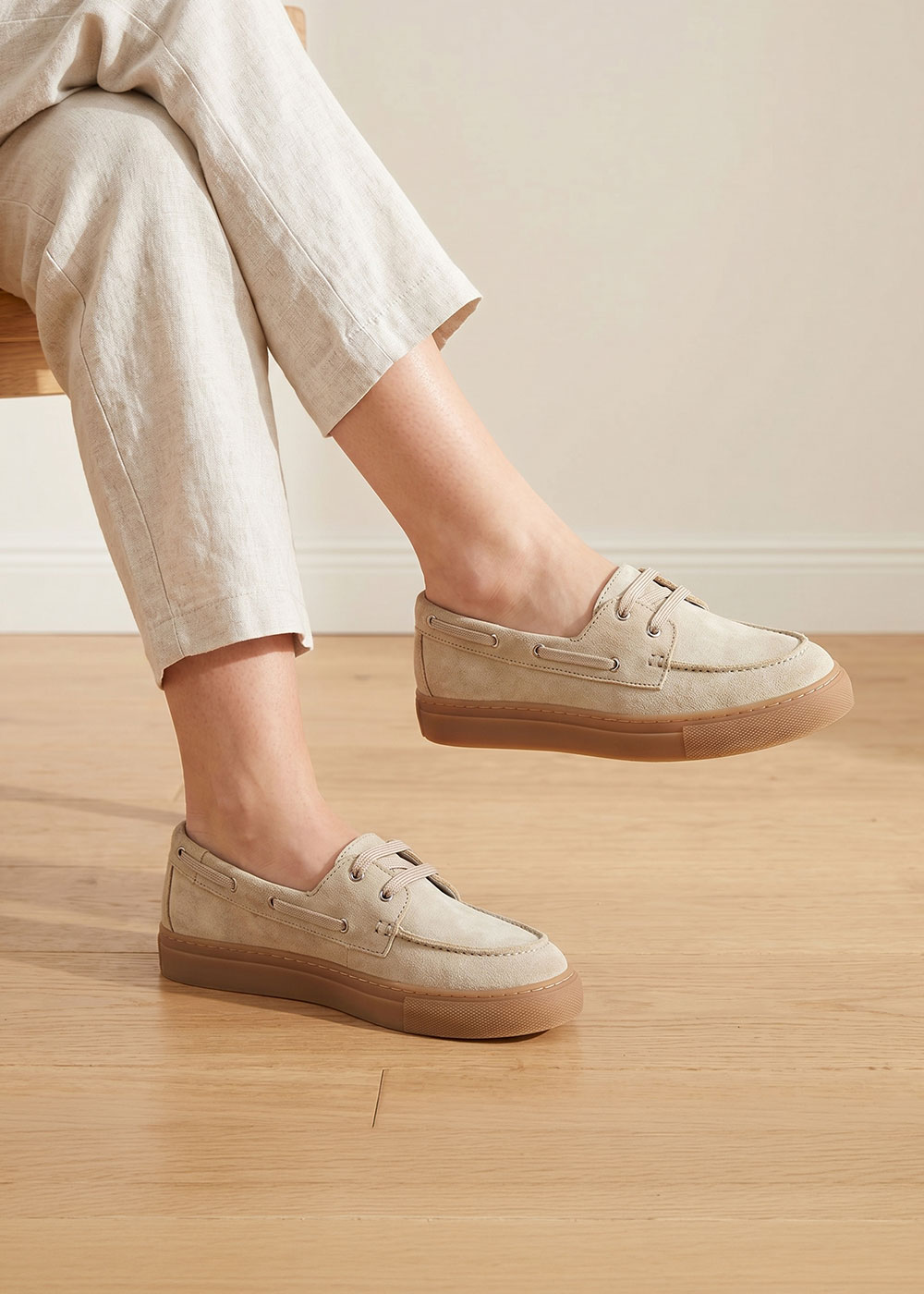 Person wearing beige suede boat shoes with gum sole and cream linen trousers, photographed on a light wooden floor