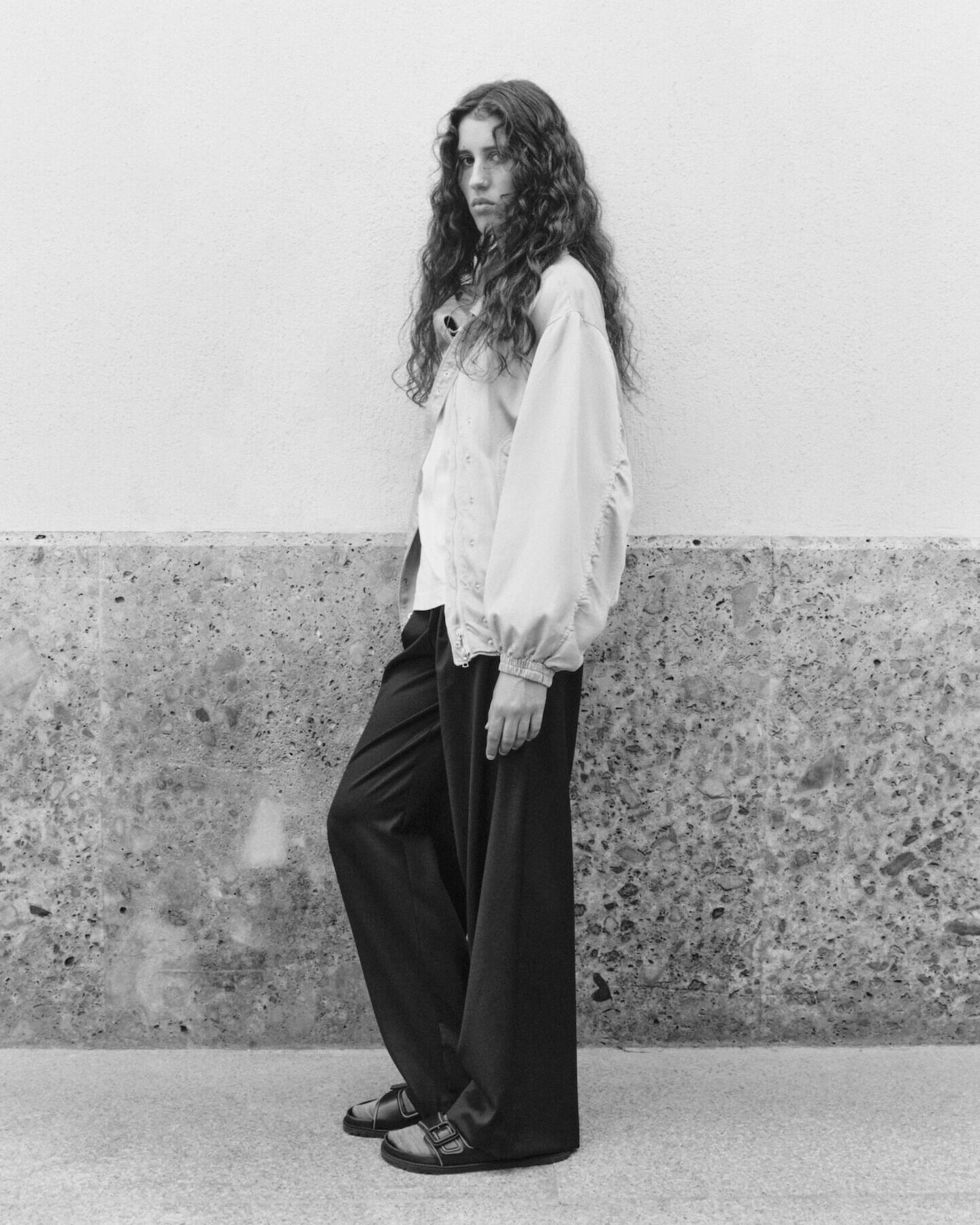 Black-and-white full-length portrait of a young woman with long curly hair wearing an oversized jacket and wide trousers, standing against a textured wall