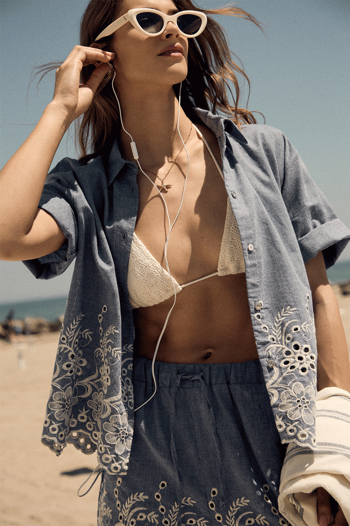 Woman at beach wearing sunglasses and earphones in a two-panel seaside fashion shot: embroidered shirt over bikini and a white flowing dress