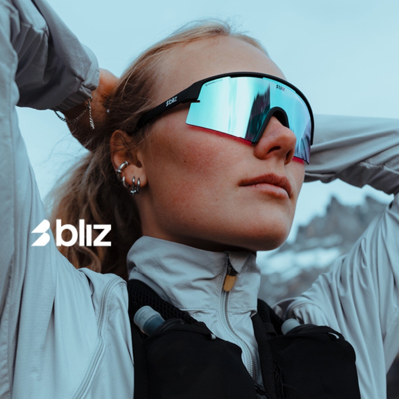 Athlete wearing mirrored Bliz sports sunglasses and light jacket with hydration pack, looking up against a snowy mountain backdrop