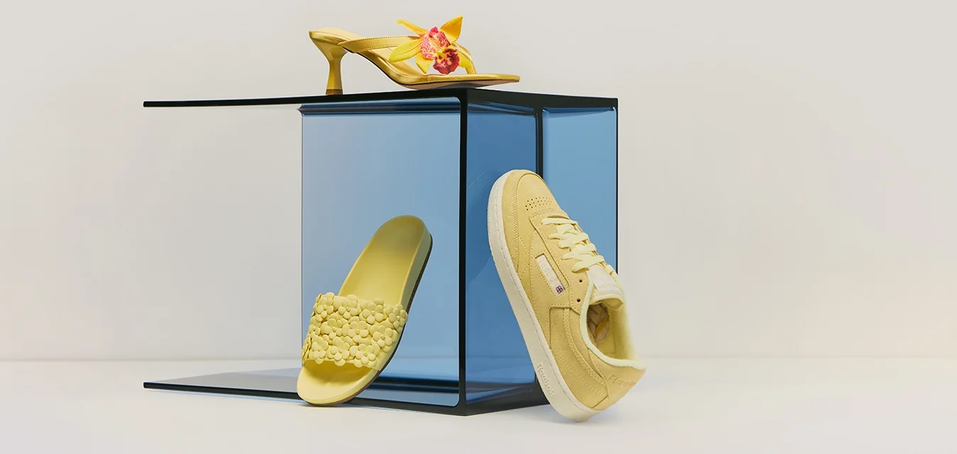 Tre scarpe gialle—un tacco alto floreale, una ciabatta con texture e una sneaker casual—esposte su e intorno a un cubo di vetro blu.
