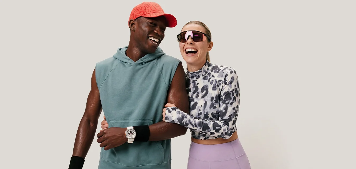 Uomo con felpa senza maniche e cappellino rosso sorride con donna con top fantasia e occhiali da sole mentre camminano braccio nella braccio su sfondo uniforme.