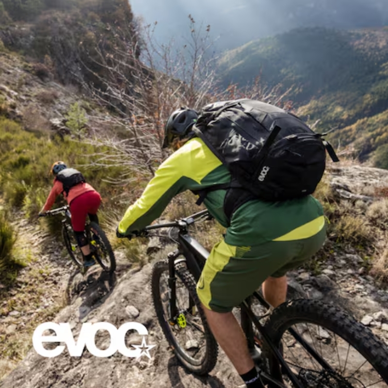 Two mountain bikers wearing helmets and backpacks descending a rocky mountain trail through grassy slopes and distant valleys