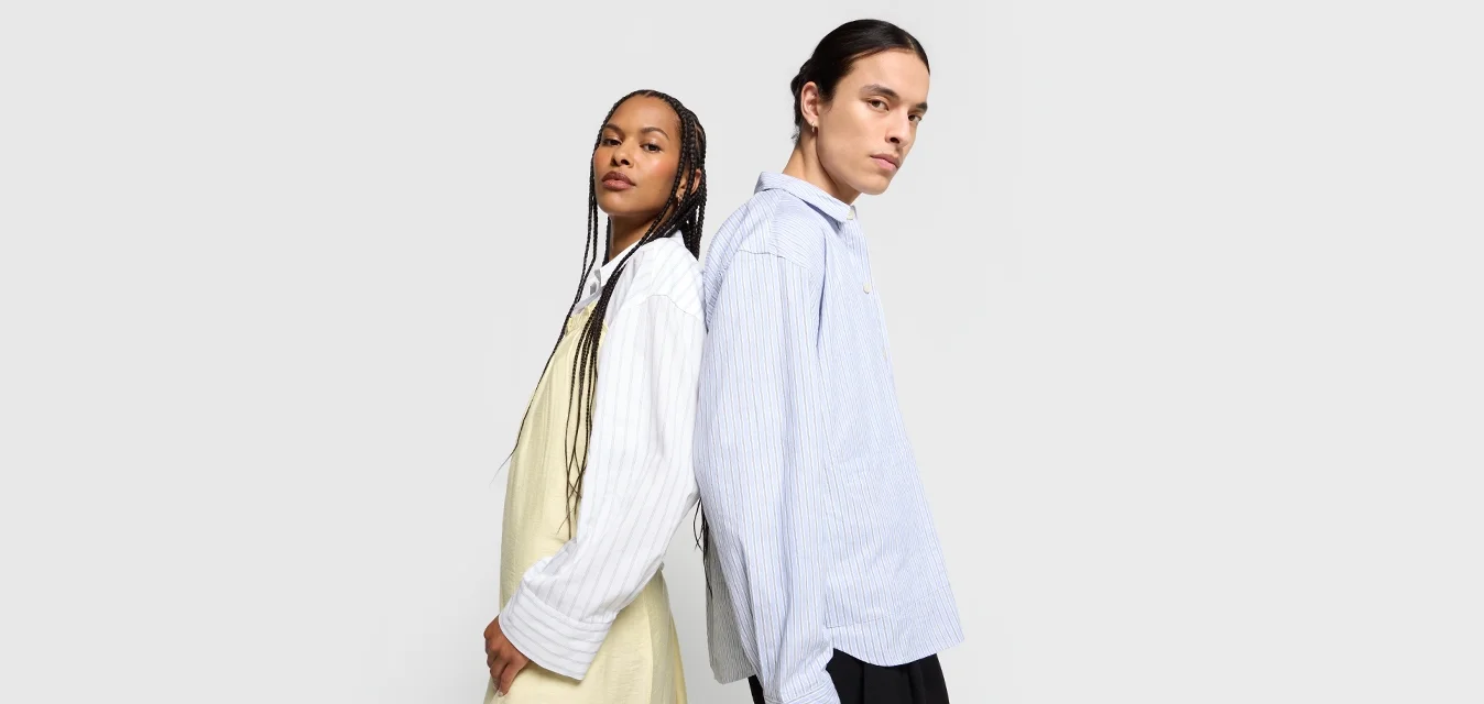 Camicia a righe blu oversize indossata da un modello; l'altro indossa un vestito giallo chiaro fluente. Entrambi hanno lunghi capelli e sono posati schiena contro schiena.