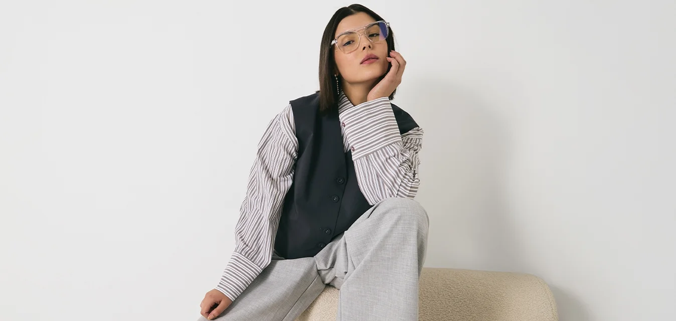 Pantaloni grigi oversize, gilet nero su una camicia a righe, occhiali con montatura blu chiaro. Seduto su un divano beige testurizzato contro una parete bianca.