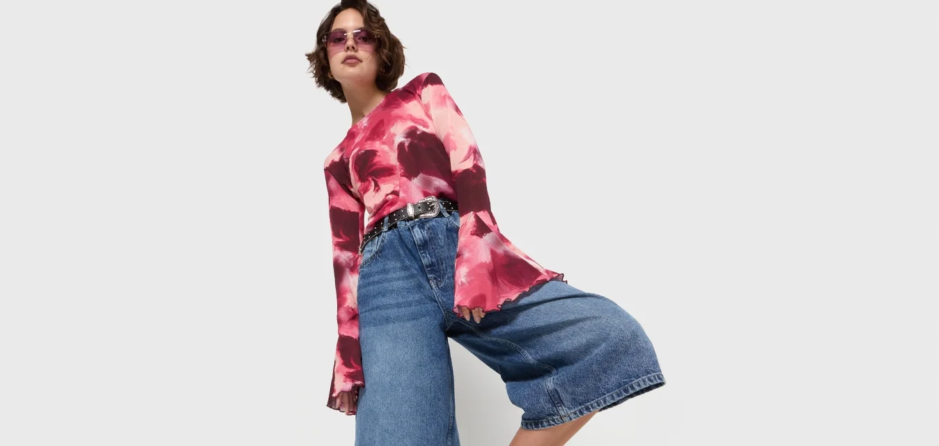Camicetta a maniche lunghe in rosso e rosa tie-dye, culotte in denim blu e sandali neri con dettagli in fibbia. Uno sfondo semplice mette in risalto l'outfit.