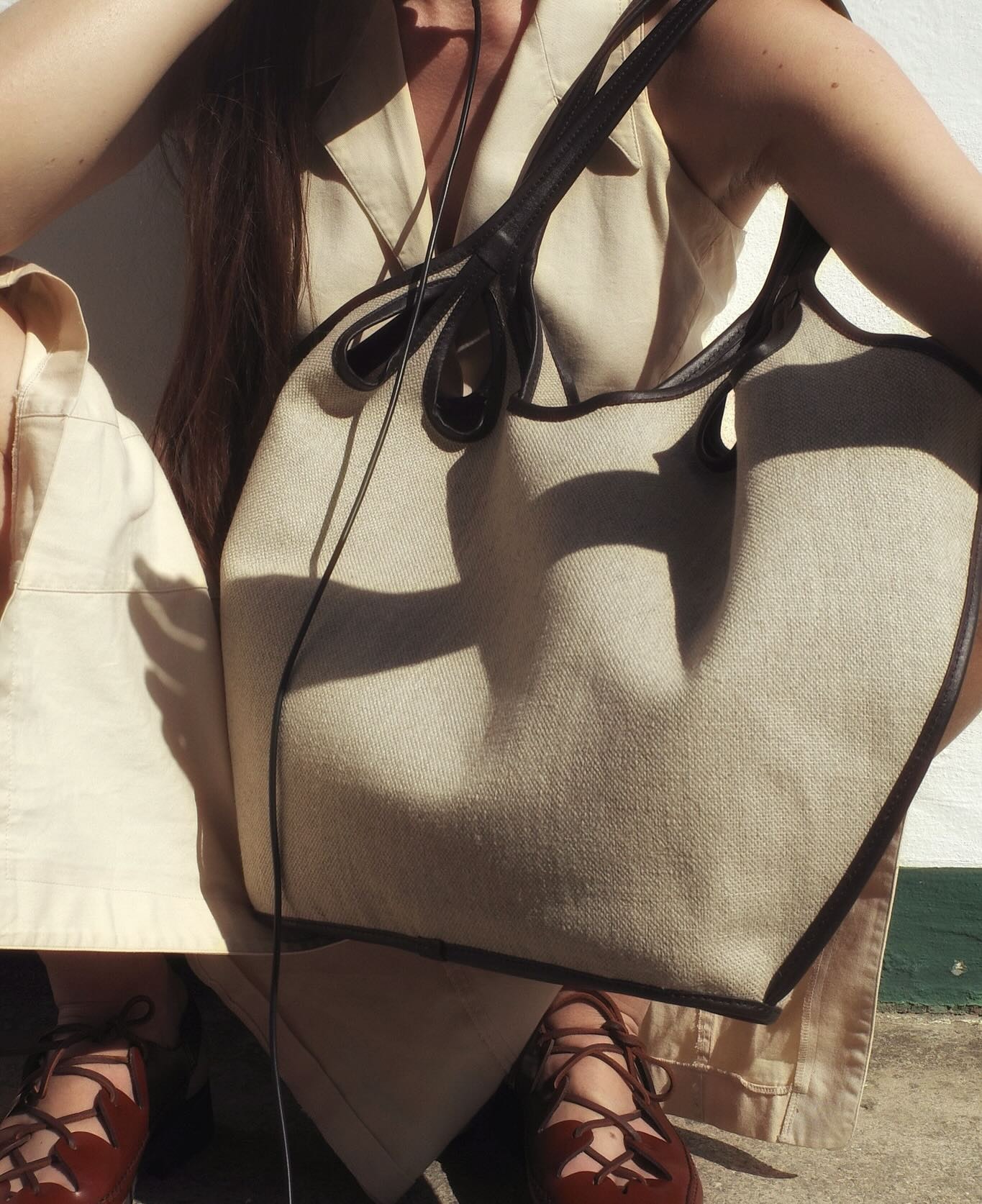 Close-up of a beige canvas tote bag with dark leather trim held against a beige summer outfit and brown lace-up shoes