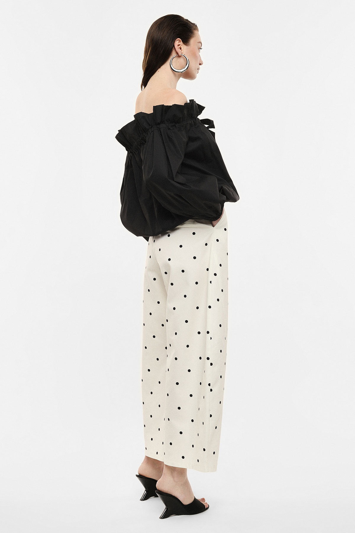 Side view of a model in an off-shoulder black ruffled blouse, cream wide-leg polka-dot trousers and black mule heels