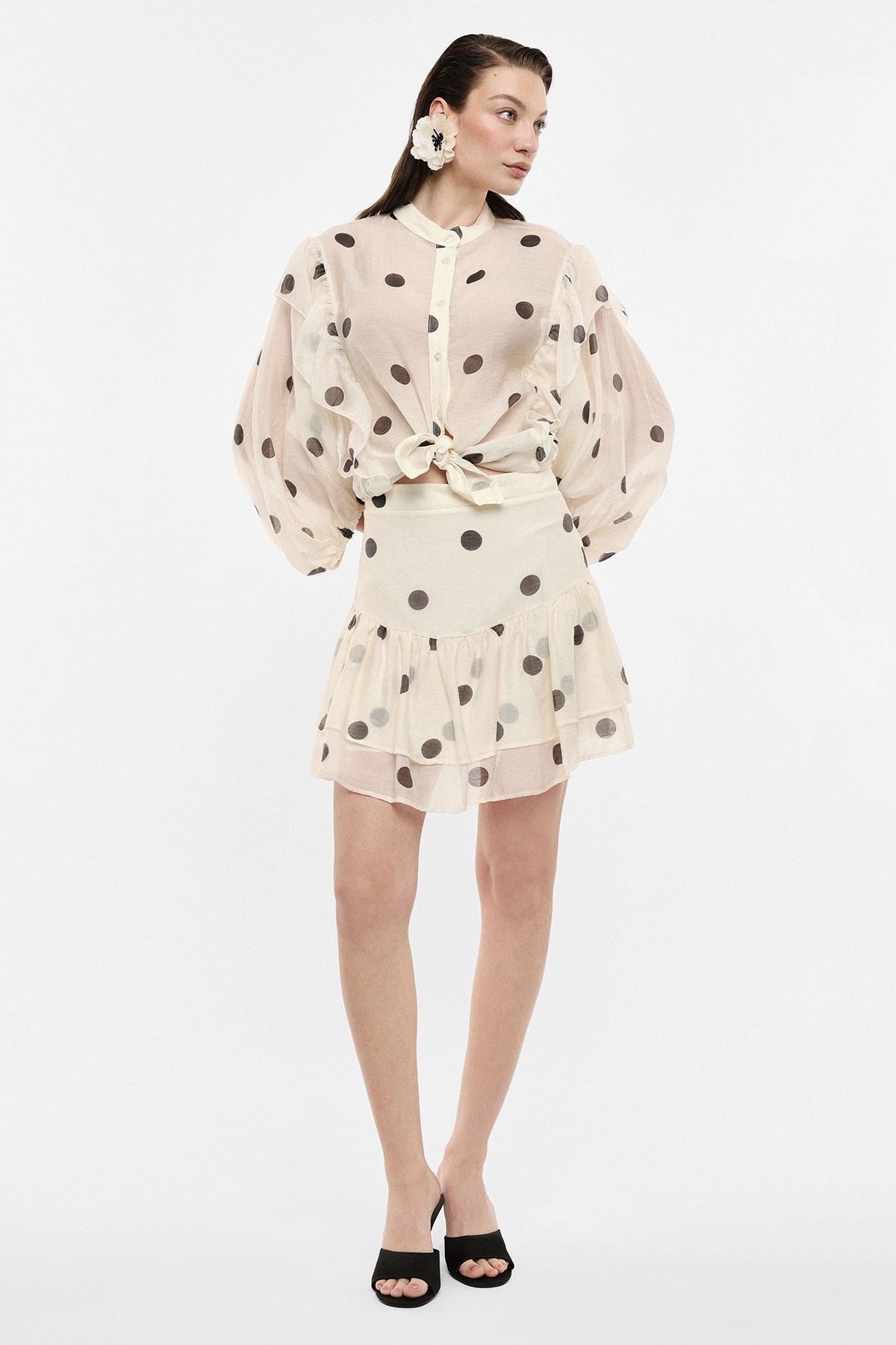 Woman modelling cream sheer polka-dot blouse tied at the waist with matching ruffled mini skirt, floral earring and black heels
