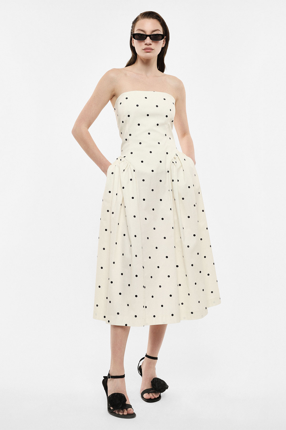 Woman wearing strapless cream midi dress with black polka dots, side pockets, black ankle-strap heels and sunglasses
