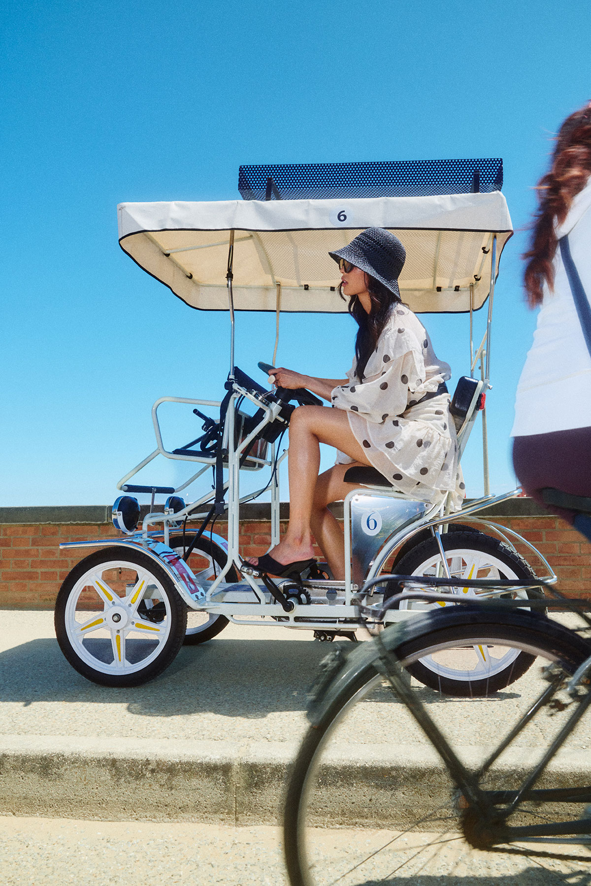 Woman in polka-dot dress and sun hat pedalling a four-wheel surrey bike along a sunny seafront promenade under a clear blue sky