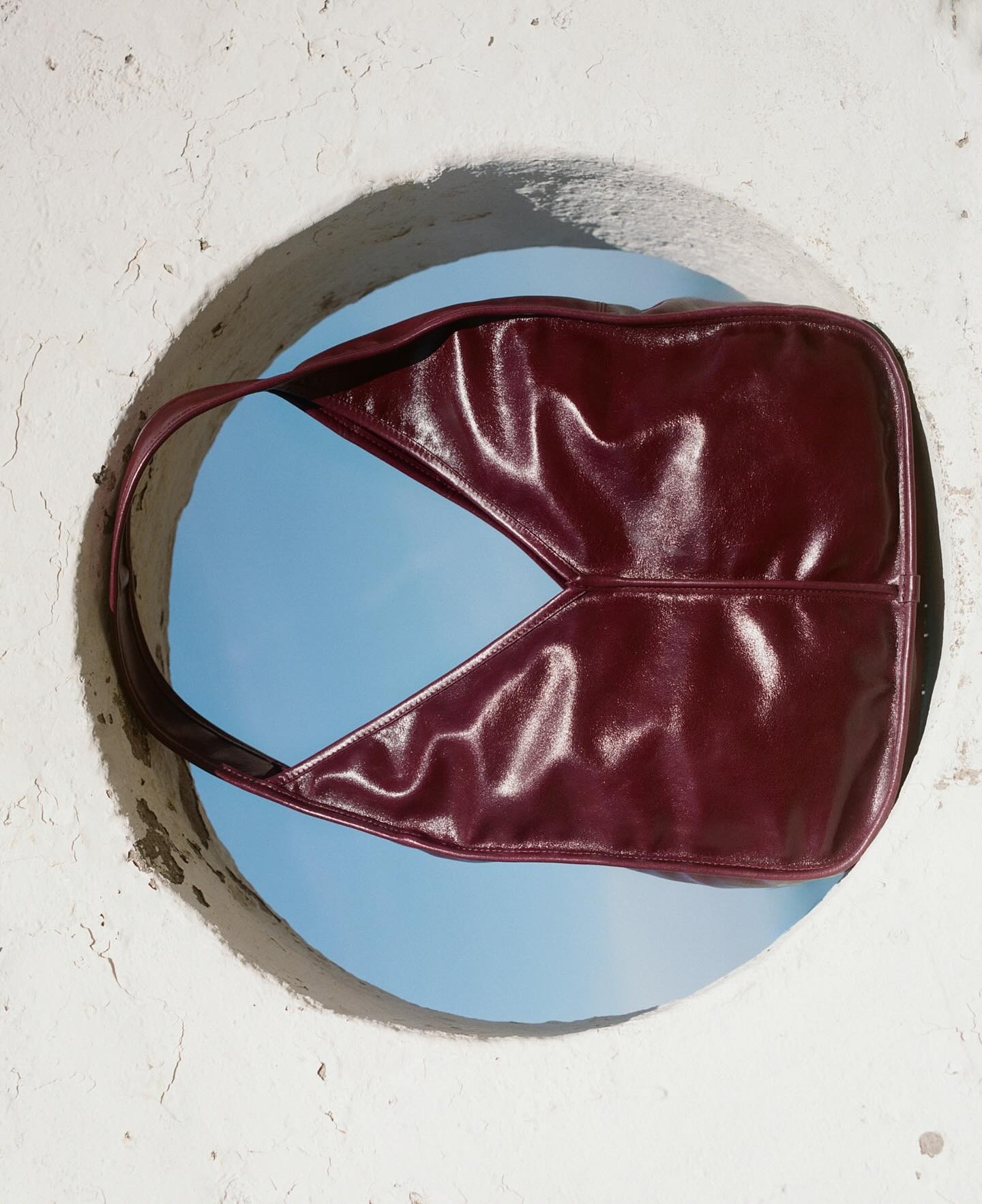 Maroon leather shoulder bag suspended in a circular white wall opening against a blue sky