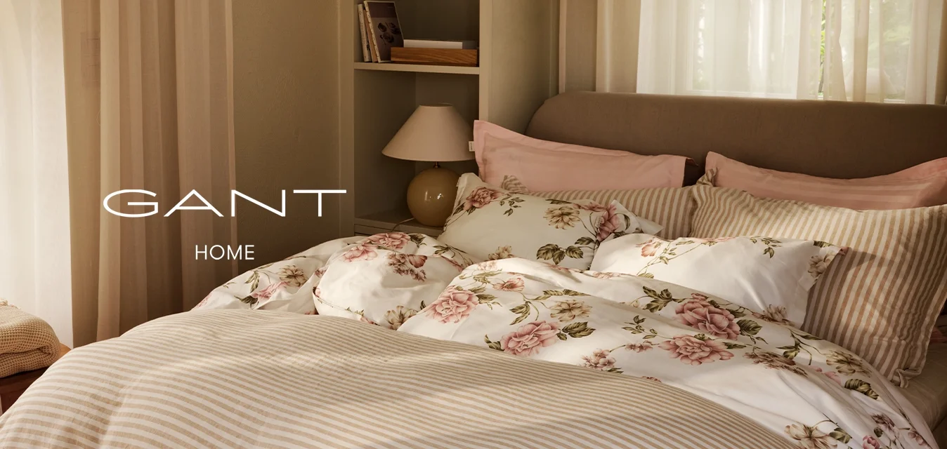 Camera da letto soleggiata con un letto ben rifatto, con biancheria da letto a righe e floreale, tende leggere, una lampada da comodino e uno sgabello piccolo con coperte piegate.
