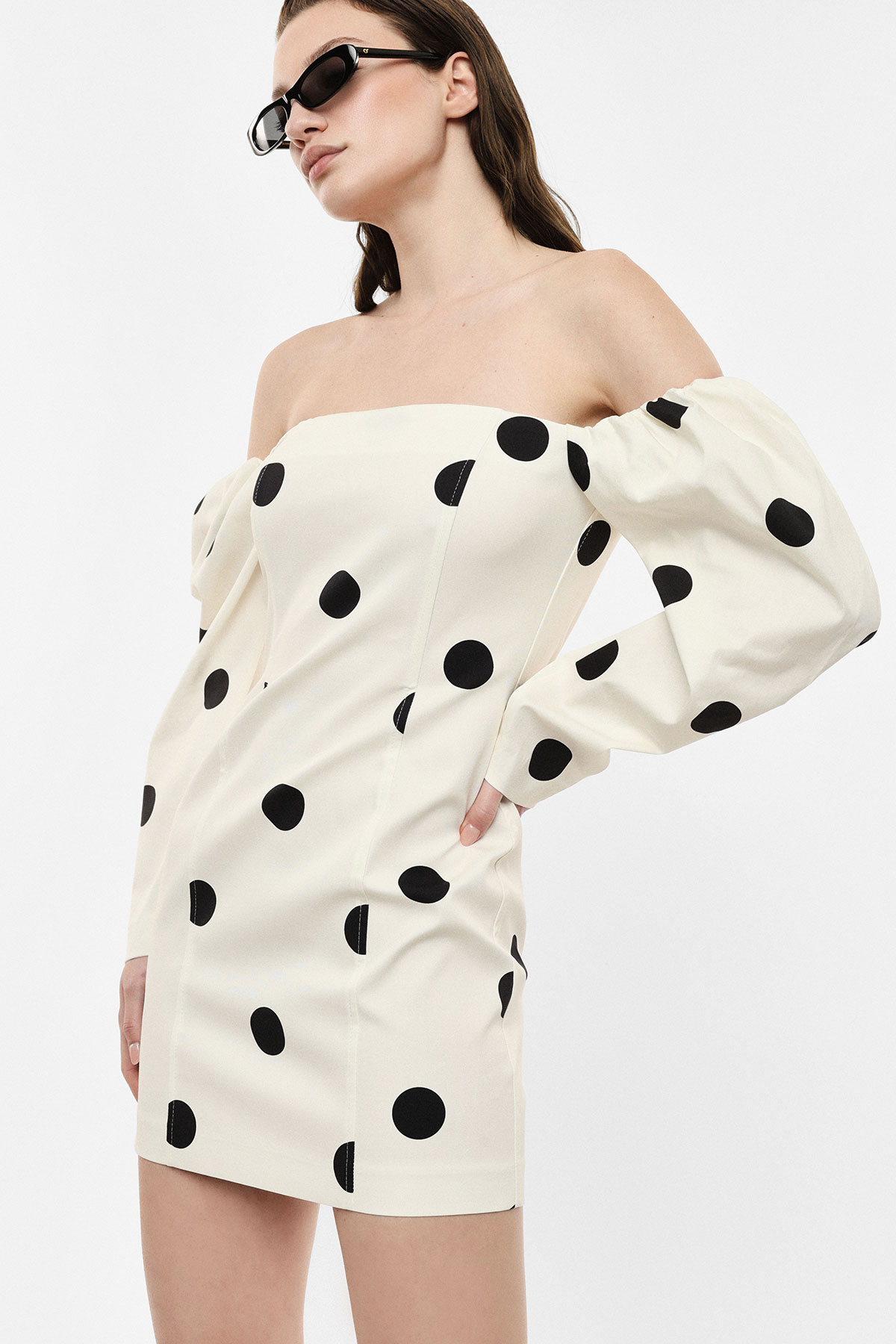 Woman wearing cream off-the-shoulder mini dress with black polka dots and voluminous puff sleeves, accessorised with black sunglasses