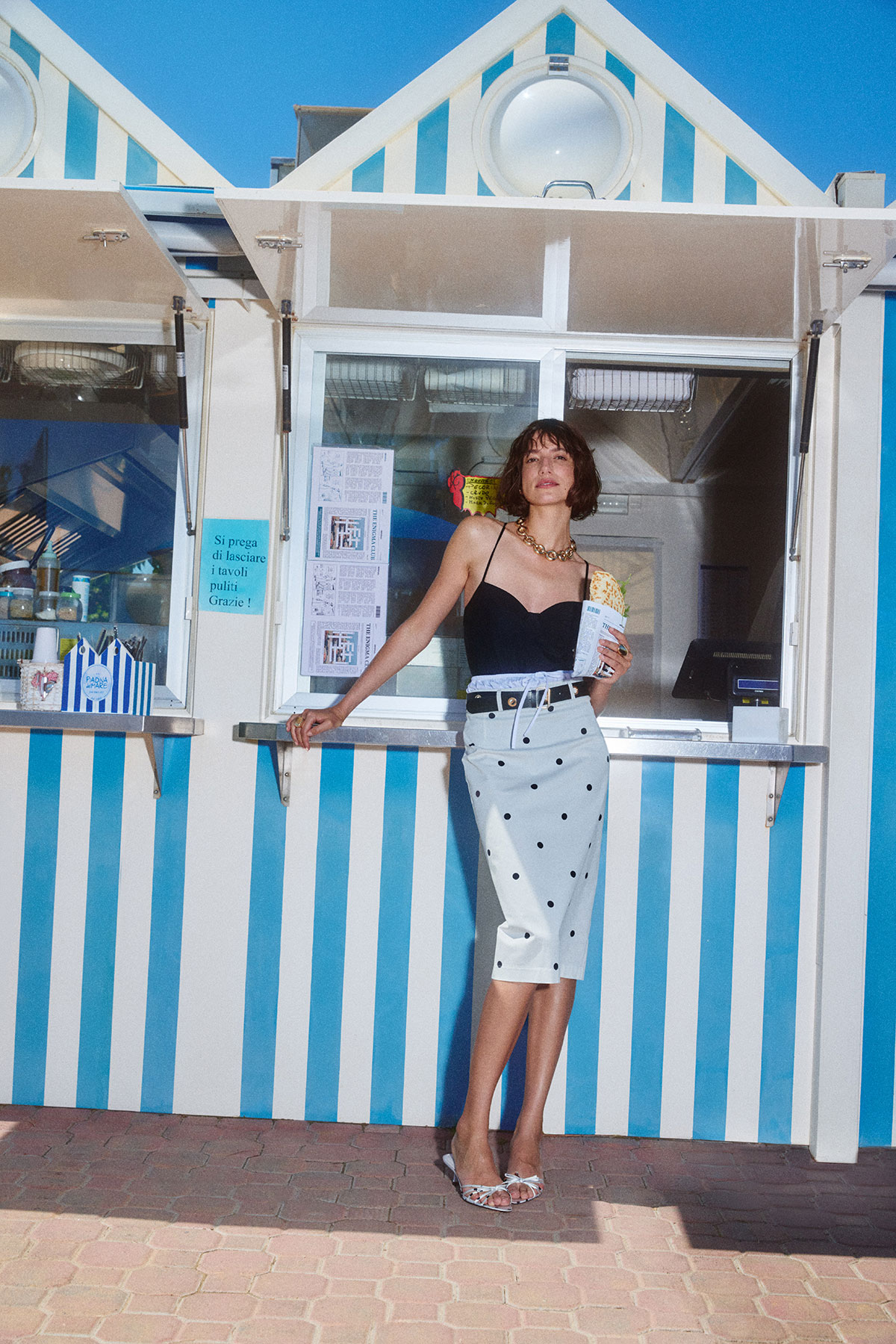 Woman in black camisole and polka-dot skirt standing at a blue-and-white striped beach kiosk holding a snack and menu