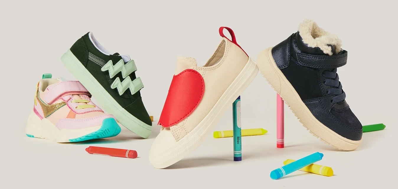 Quattro scarpe colorate per bambini in equilibrio su dei pastelli colorati, con altri pastelli sparsi su uno sfondo bianco.