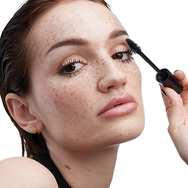 Primo piano di una modella con lentiggini che applica il mascara nero sulle ciglia superiori con uno scovolino YSL LASH CLASH.