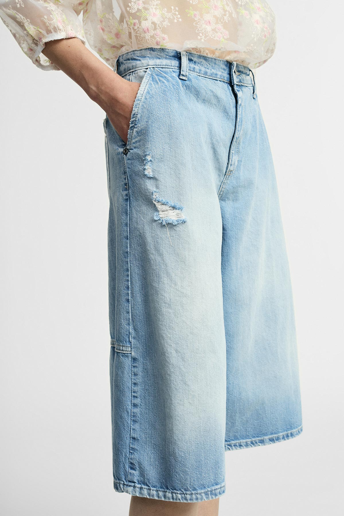 High-waist light-blue wide-leg knee-length distressed denim culotte shorts with side pockets, model wearing sheer floral blouse