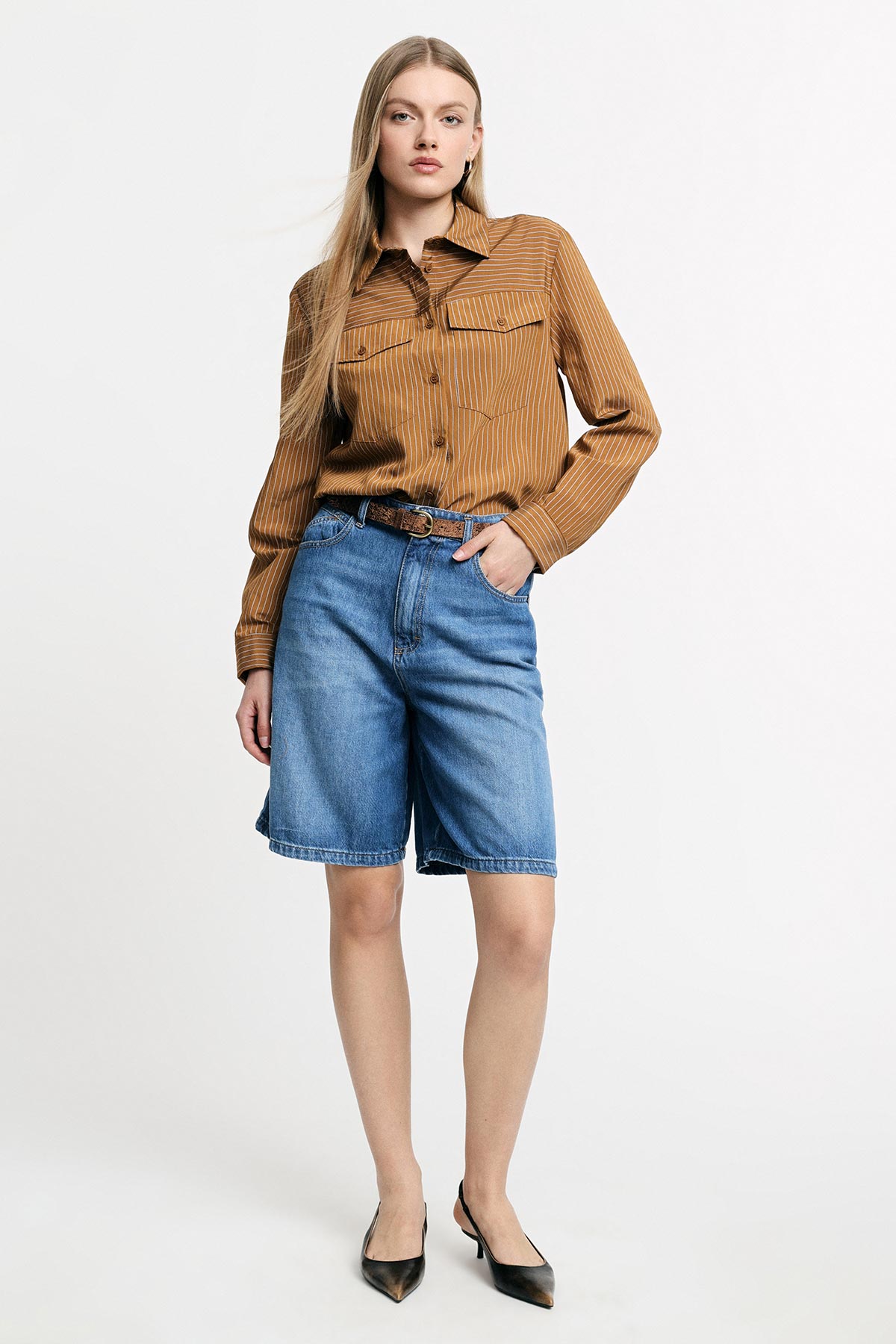Model wearing brown striped shirt tucked into blue denim bermuda shorts with belt and black heels