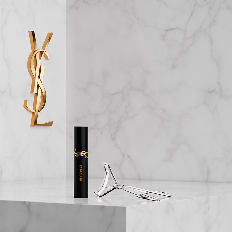 Un MINI LASH CLASH MASCARA e un PIEGACIGLIA YSL argento su un piedistallo di marmo bianco accanto al logo Cassandre dorato.