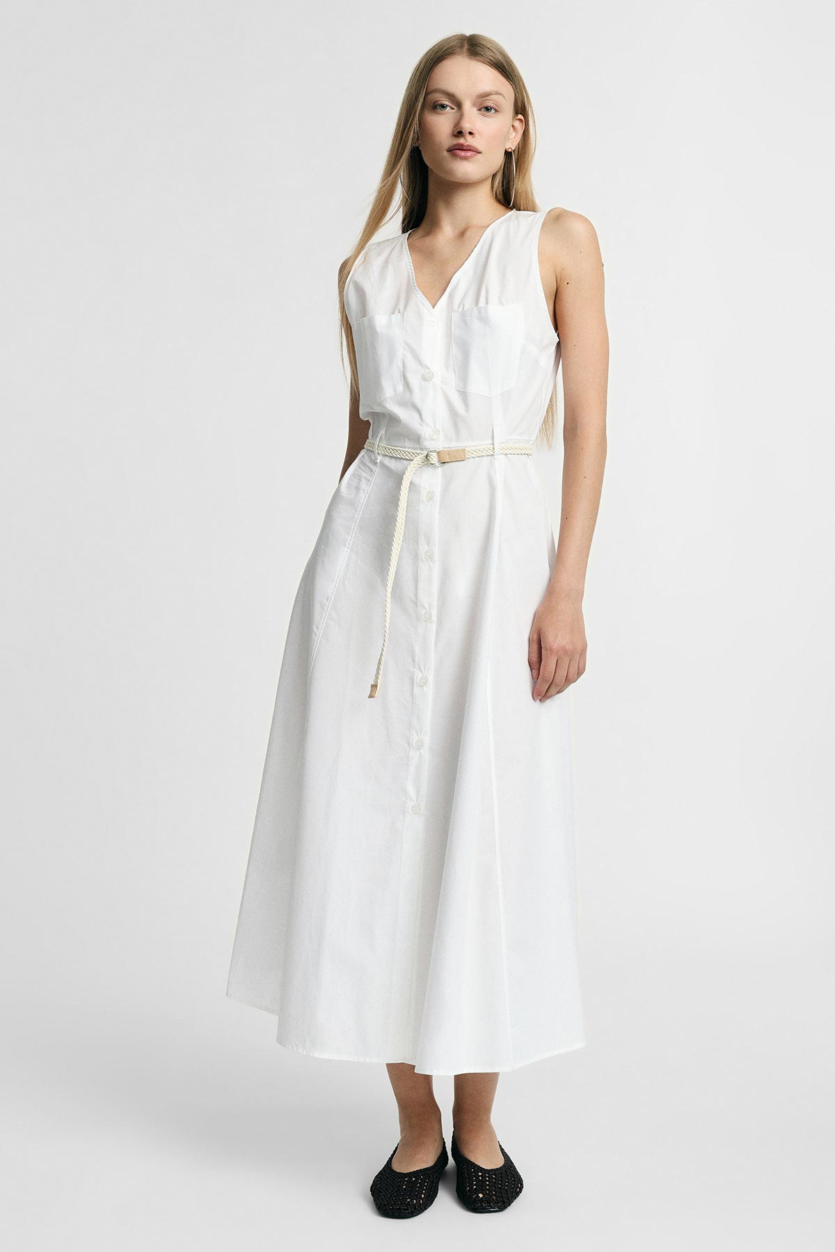 Woman wearing sleeveless white button-front midi dress with thin braided belt and black flats on plain studio background