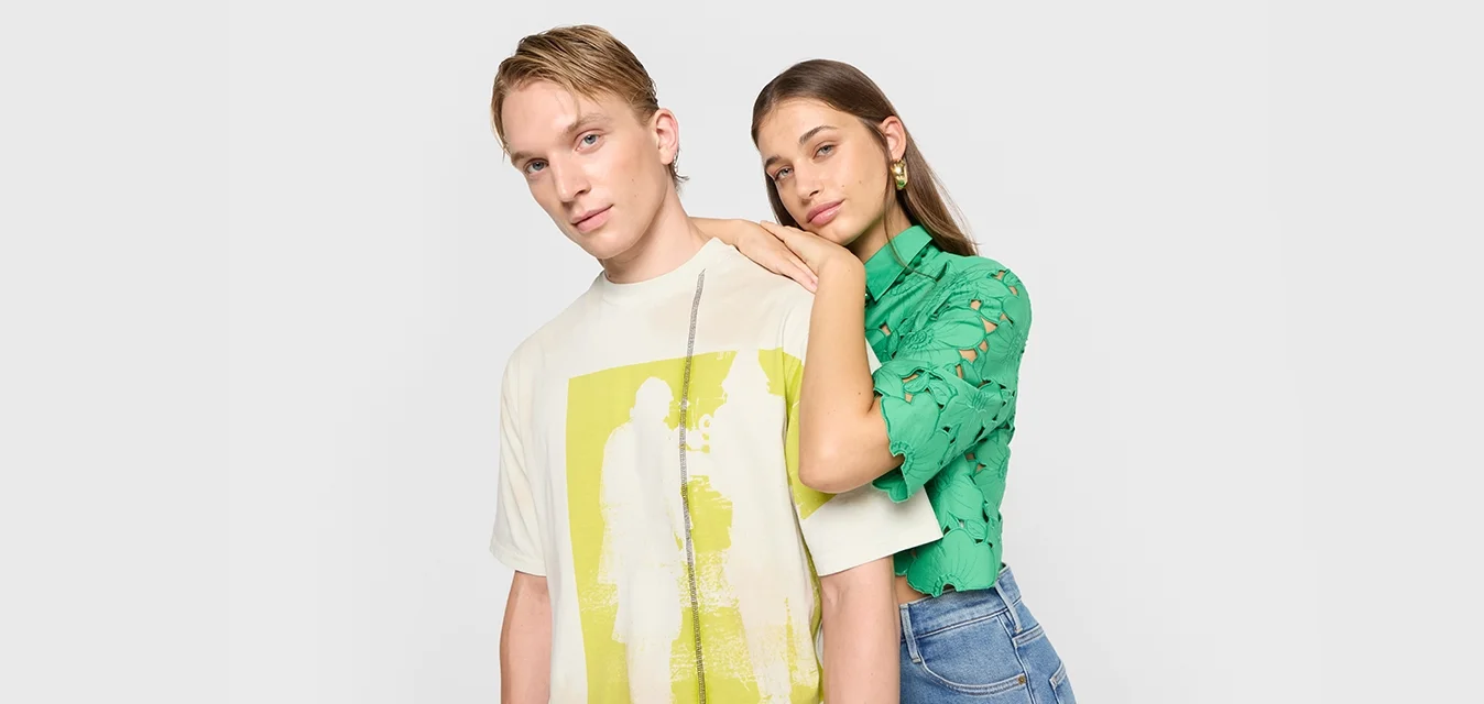T-shirt grafica color crema con stampa gialla; top verde a maniche corte con intagli floreali. Entrambi abbinati a jeans blu.