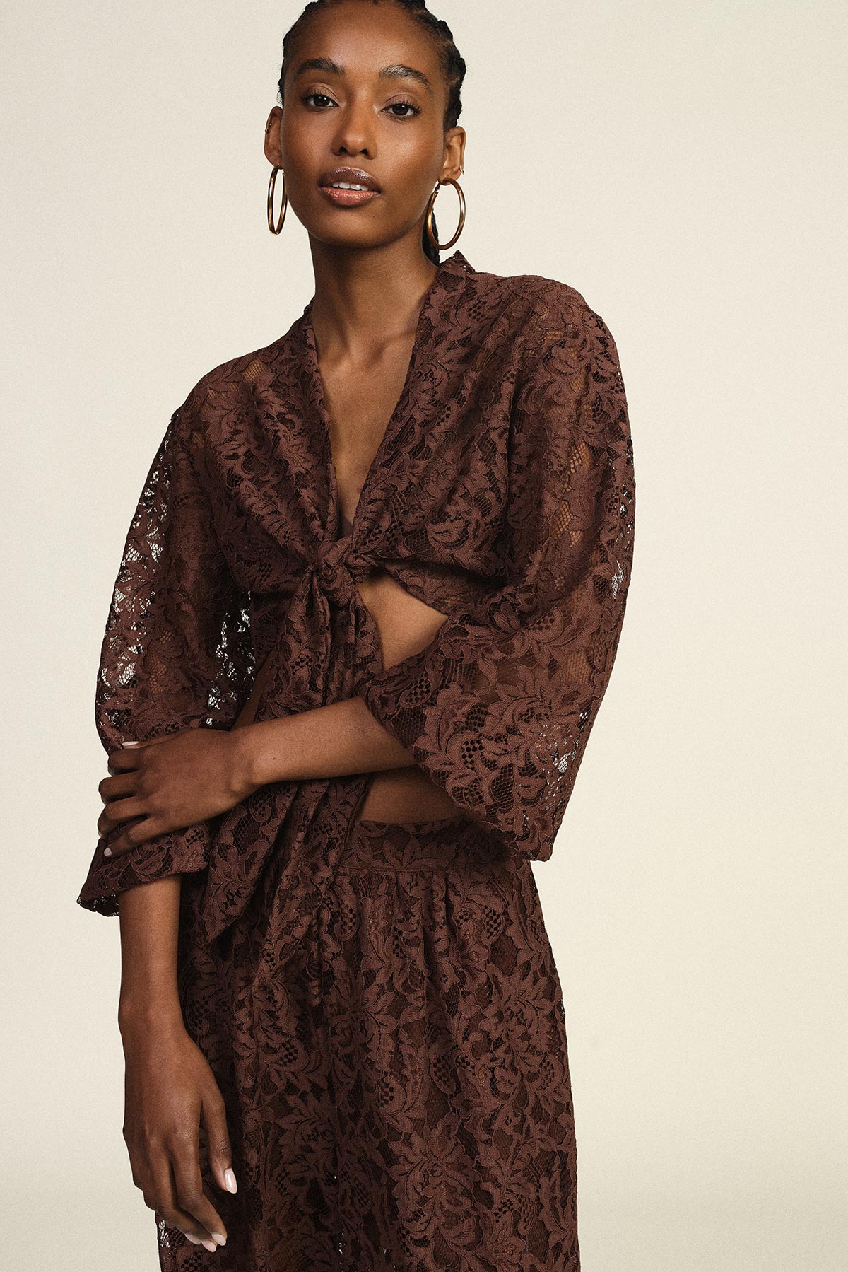 Model wearing brown lace tie-front co-ord with sheer three-quarter sleeves and matching wide-leg trousers, gold hoop earrings