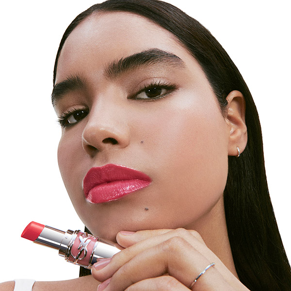 Primo piano di una modella con labbra magenta intense, che tiene un rossetto YSL LOVESHINE. Espressione potente e audace.