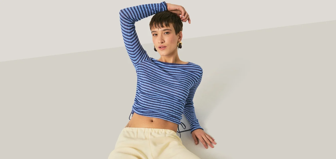 Giovane che si rilassa con un braccio dietro la testa, indossando una camicia a righe blu a maniche lunghe e pantaloni beige chiaro, su uno sfondo semplice.