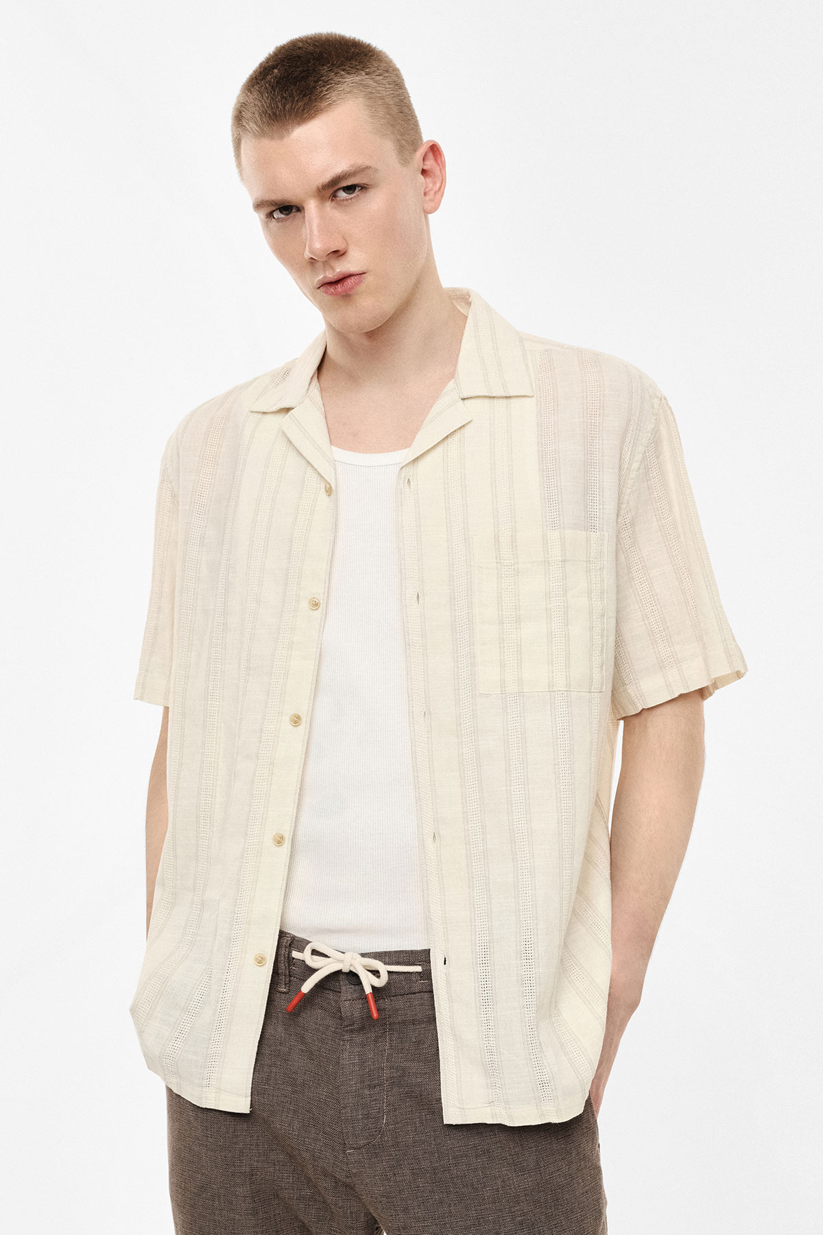 Young man wearing cream short-sleeve linen shirt over white vest and brown drawstring trousers, hands in pockets on white background
