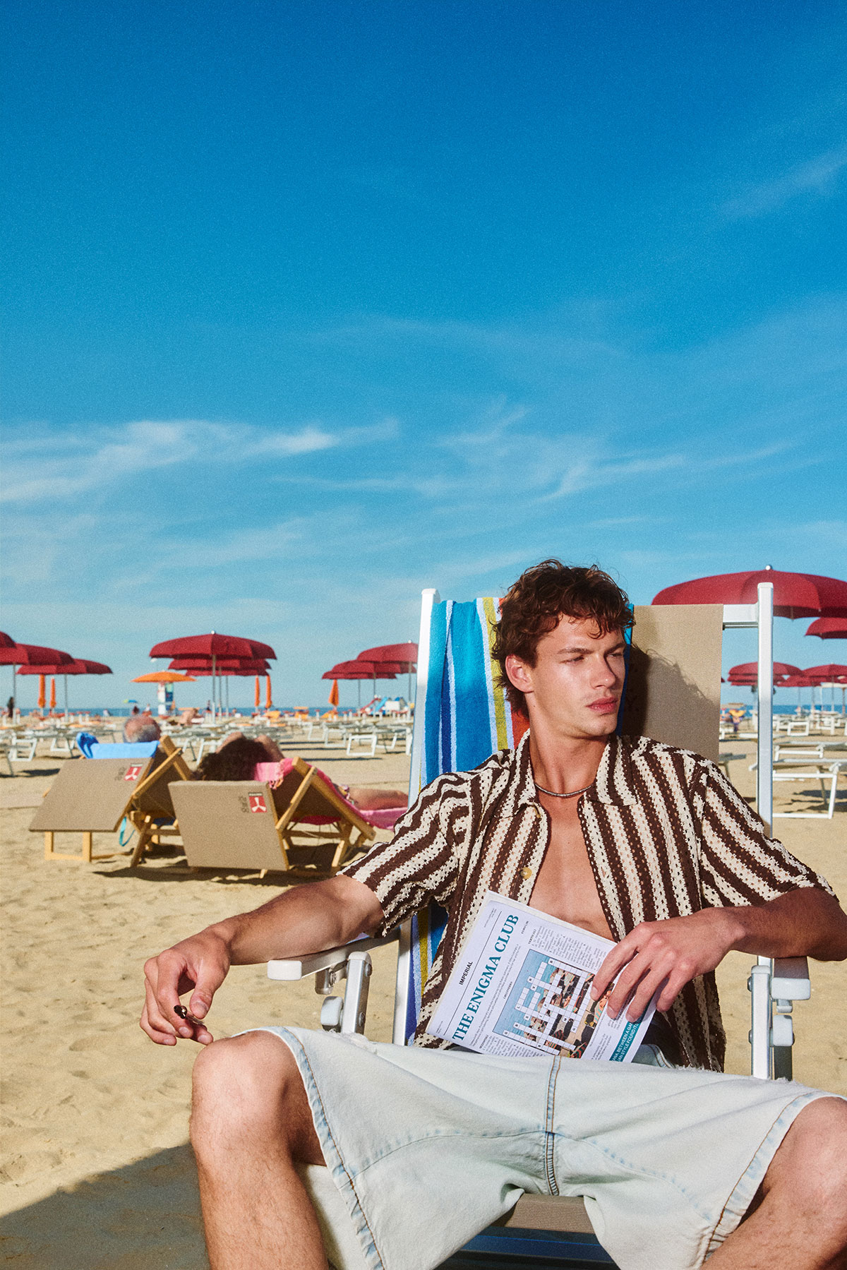 Young man in striped shirt lounging on a deck chair holding a magazine on a sunny beach with red umbrellas