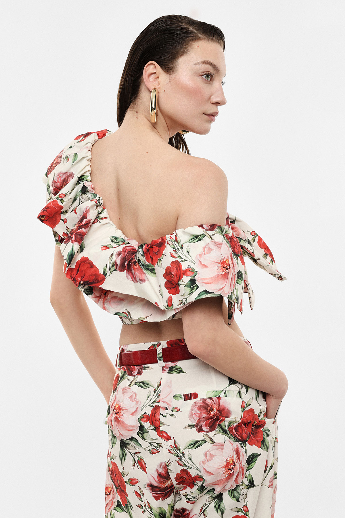 Model wearing off-shoulder floral crop top and matching high-waist trousers with red and pink roses, gold hoop earring, hands in pockets, looking right