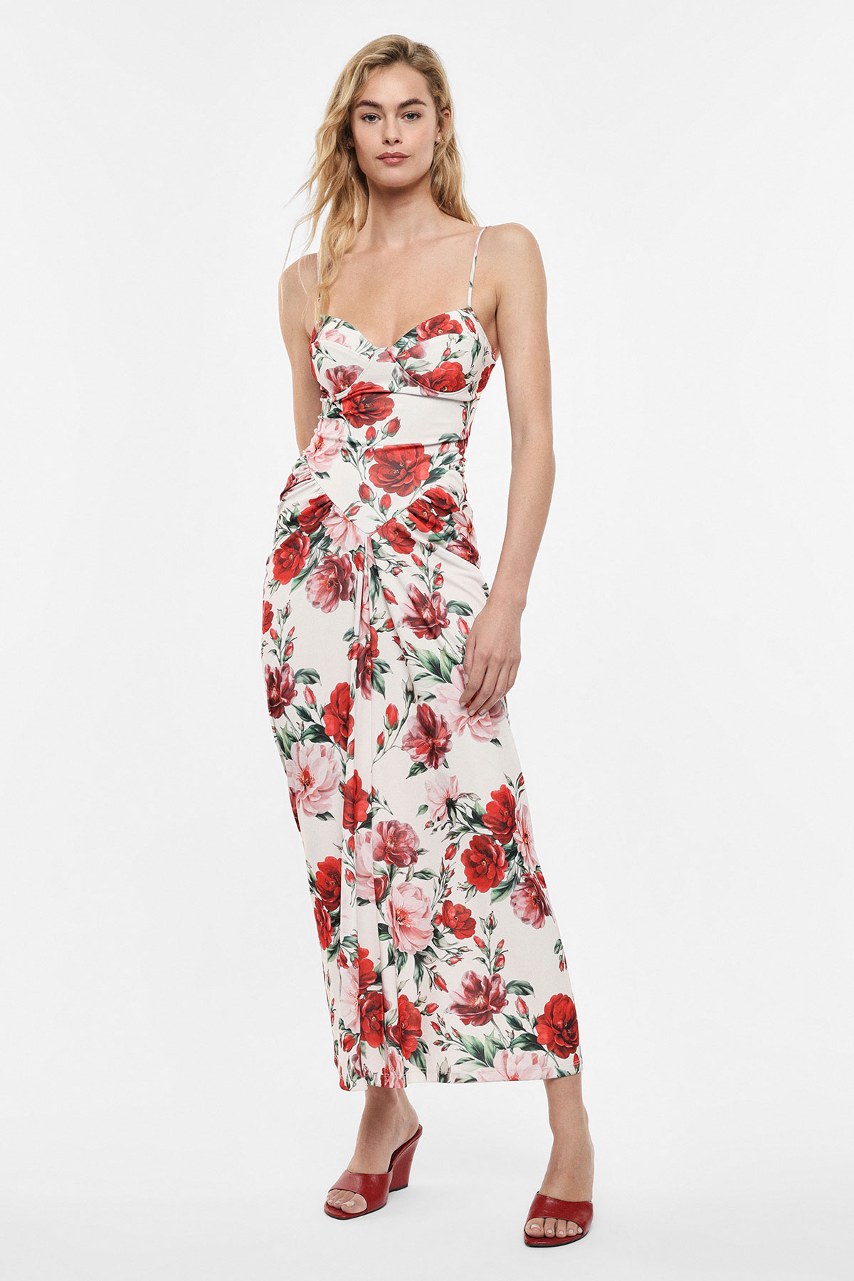 Model wearing white floral maxi dress with red rose print, spaghetti straps and ruched waist, paired with red heeled sandals