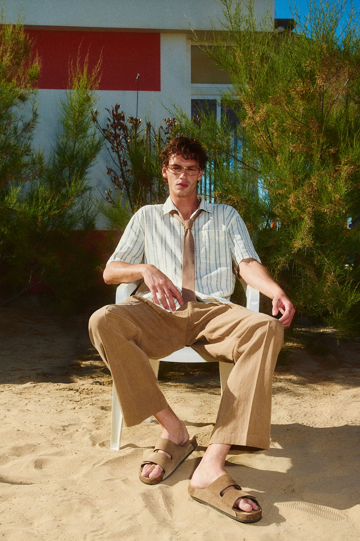 Young man in striped shirt, tie and sandals sitting on a white plastic chair on sandy beach with shrubs and a modern seaside building behind