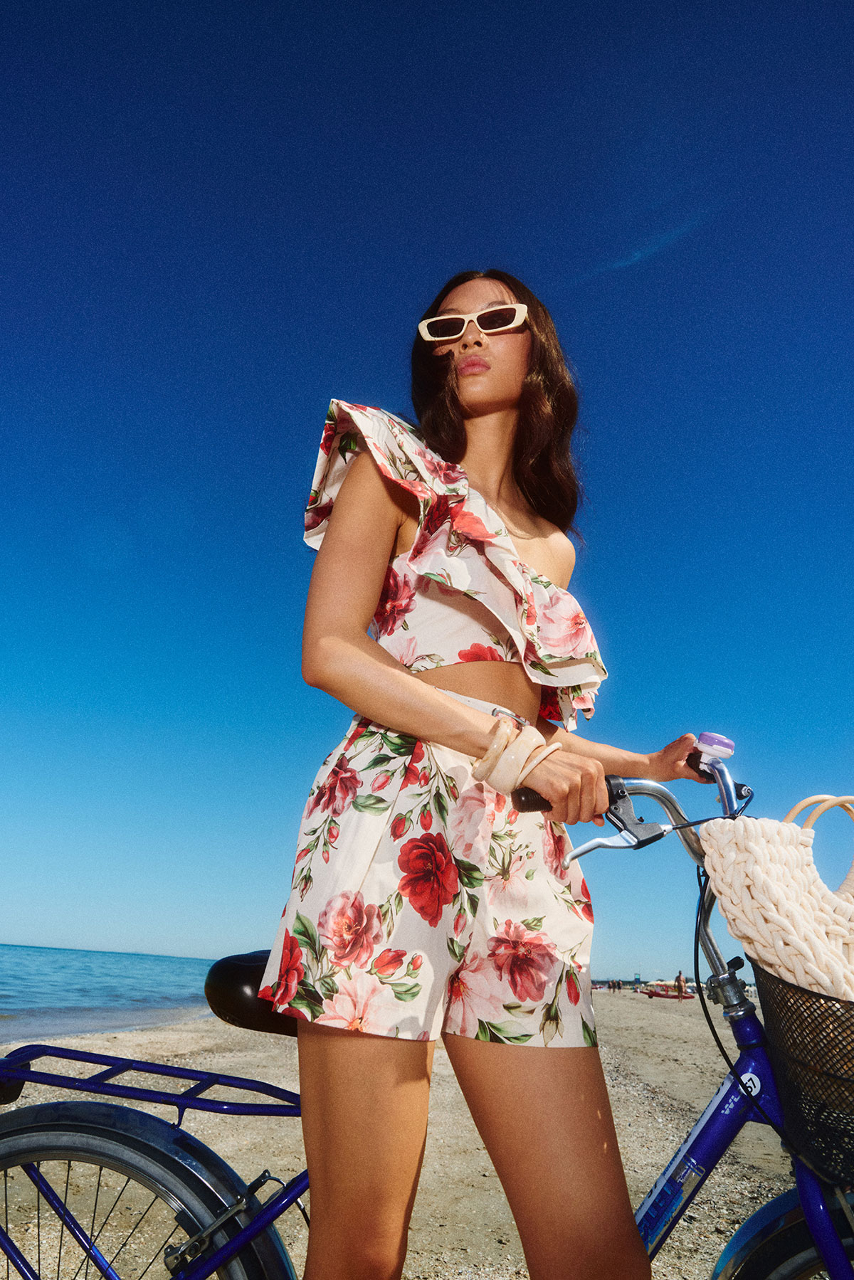 Woman in floral two-piece outfit and sunglasses standing with bicycle on a sandy beach under a clear blue sky
