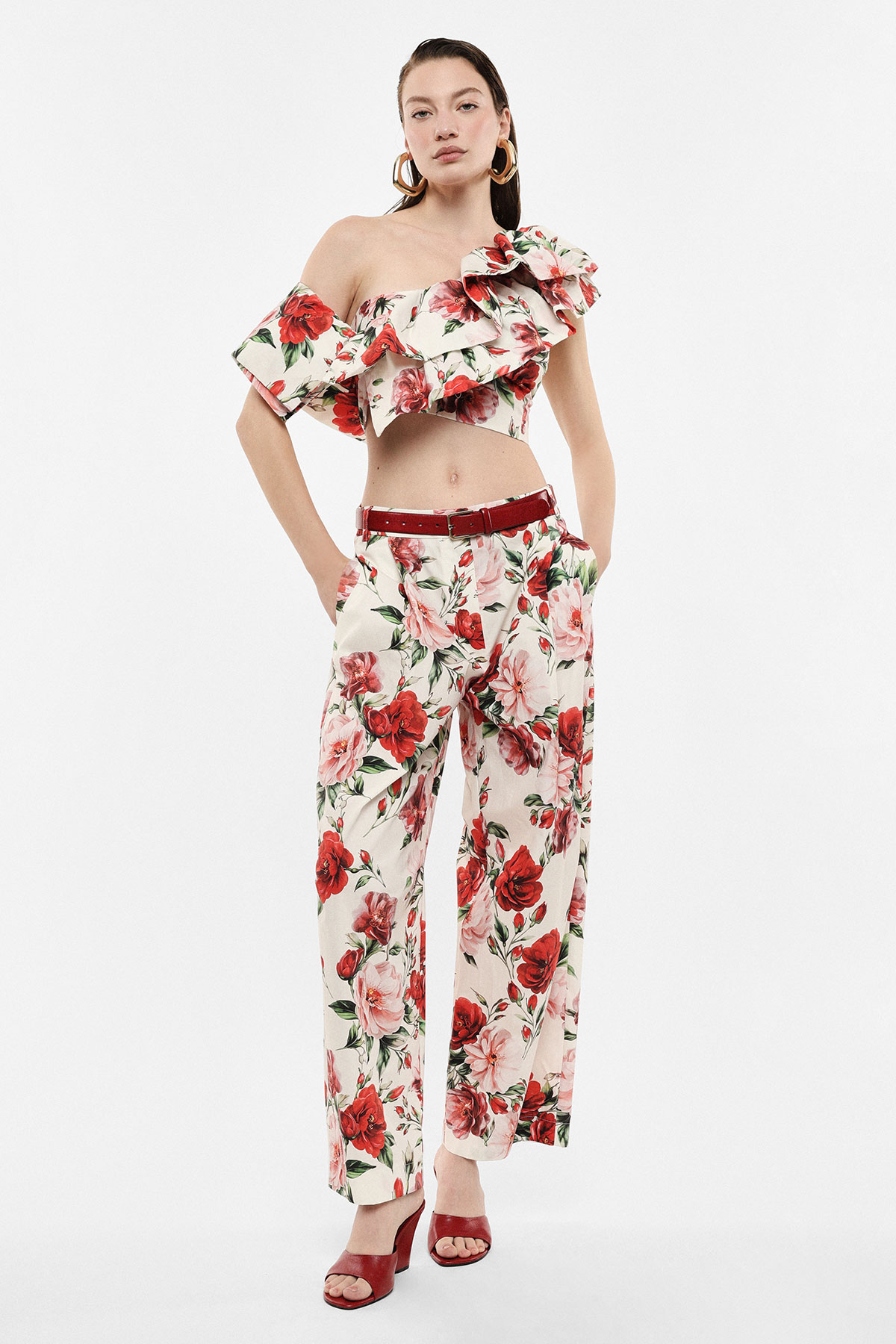 Model wearing floral off-shoulder ruffled crop top and matching wide-leg trousers with red belt and heels on white background