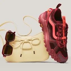 Borsa beige con dettaglio a fiocco, occhiali da sole scuri appesi alla tracolla e una scarpa sportiva rossa e rosa appoggiata alla borsa.