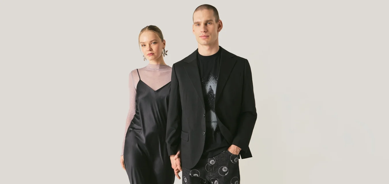 Giovane donna con vestito nero sottile e top trasparente con sandali gialli, e uomo con blazer nero e pantaloni decorati che camminano fianco a fianco.